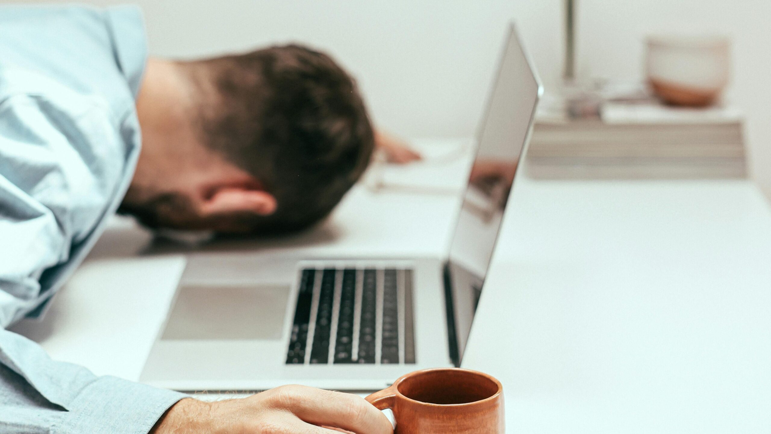 A fatigued man in an office setting asleep at his desk with a laptop and coffee mug.