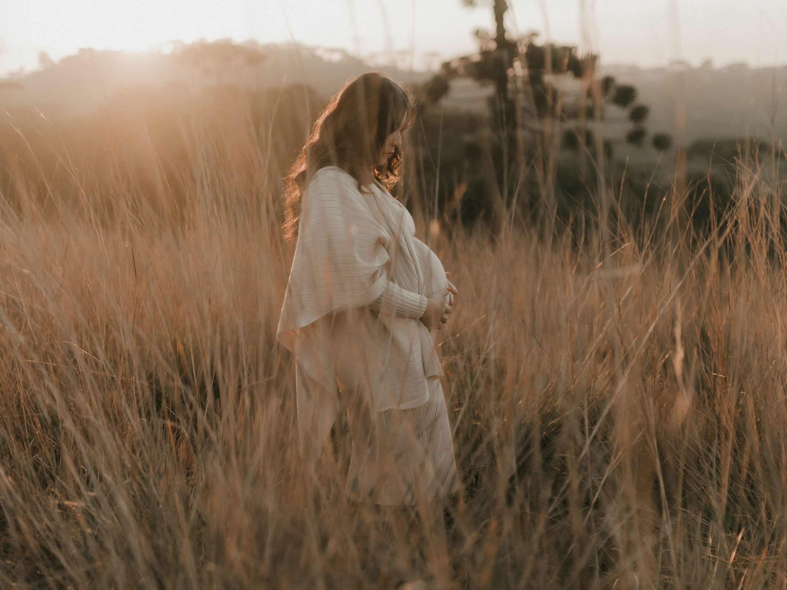 A pregnant woman stands in a sunlit field, embracing nature's tranquility.