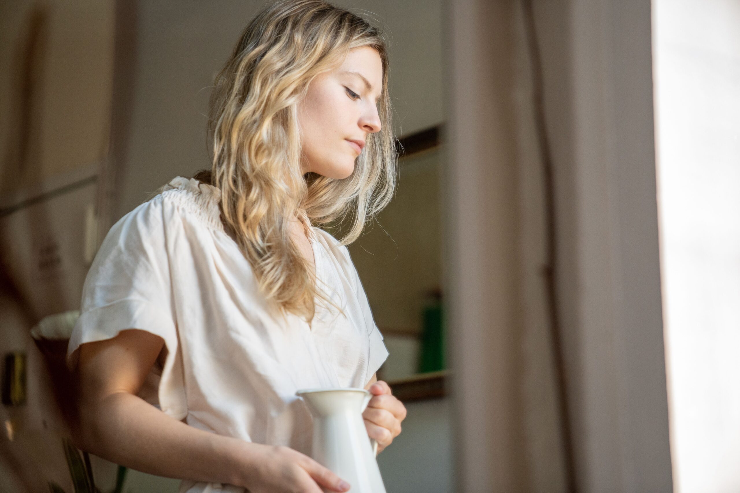 A serene portrait of a blonde woman inside holding a white pitcher, captured with natural light.